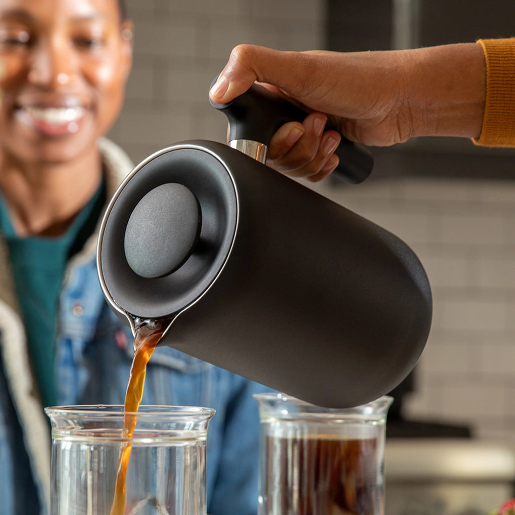 Nogen skænker kaffe fra en slank Fellow Clara French Press, Sorter med vakuumisolerede vægge i klare glaskrus. I baggrunden smiler en anden person lidt sløret i et køkken med neutrale toner.