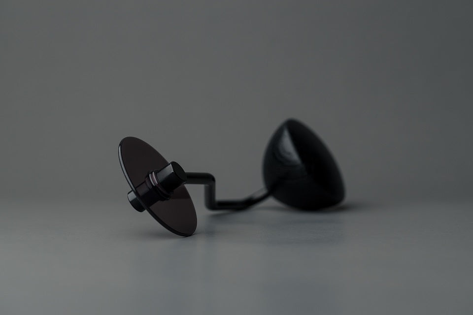 Et par sorte solbriller med ovale glas hviler på en grå overflade. Den ene linse vender opad, og armen ligner et Comandante® Sort Håndsving kværnhåndtag, hvilket skaber en minimalistisk og moderne stemning mod den glatte neutrale grå baggrund.