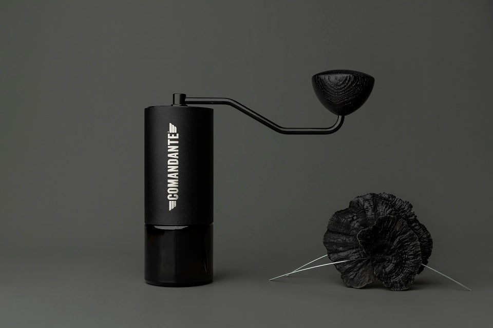 En Comandante Sort Håndsving manuel kaffekværn med en cylindrisk krop og det berømte sorte håndsving står på en mørk baggrund og fremhæver elegancen af C40 Mk3 og Mk4 kværne, med to mørke teksturerede fjer i forgrunden.