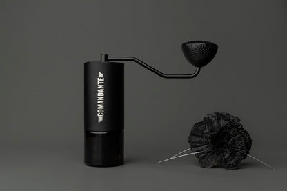 En Comandante Sort Håndsving manuel kaffekværn med en cylindrisk krop og det berømte sorte håndsving står på en mørk baggrund og fremhæver elegancen af C40 Mk3 og Mk4 kværne, med to mørke teksturerede fjer i forgrunden.