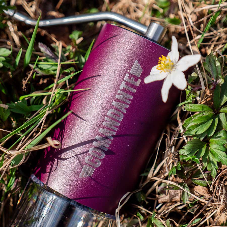 Et Comandante C40 MK4 Nitro Blade kaffekværn i Deep Purple hviler på græsset ved siden af en hvid blomst og grønne blade, og sollyset fremhæver den metalliske finish.