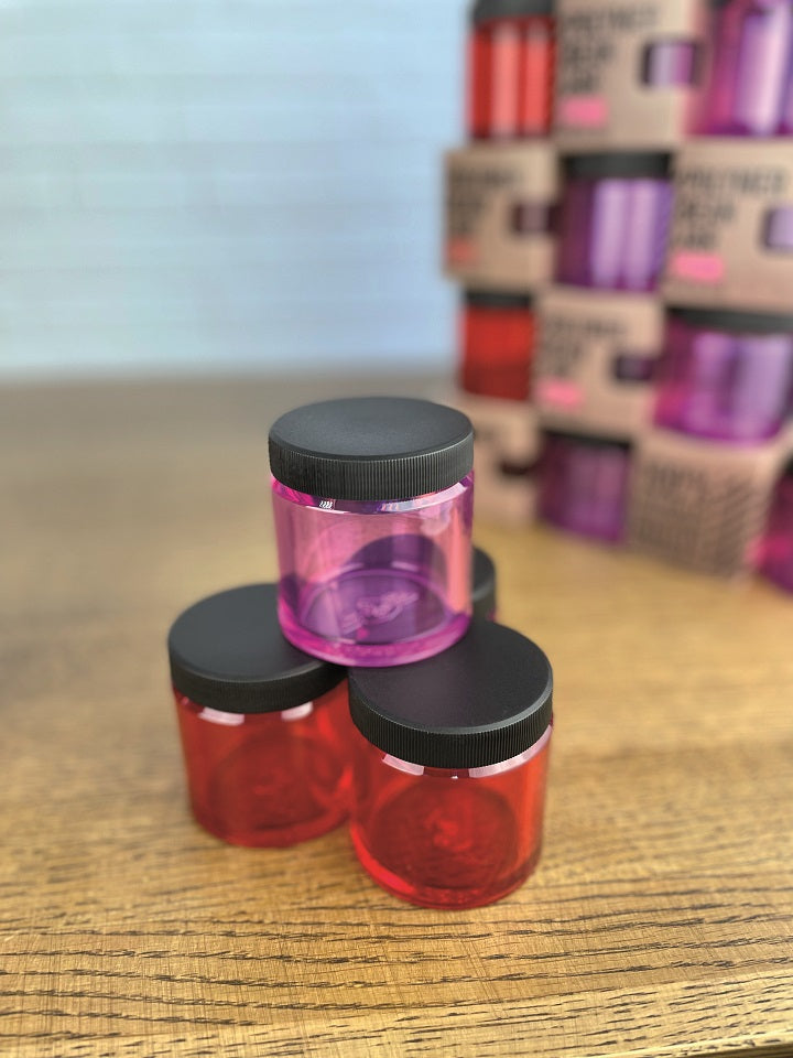 Tre farverige krukker, en pink og to røde, med sorte låg er stablet på en træoverflade. Blandt dem skiller en Comandante Polymer Bean Jar med låg i Rød sig ud. Flere krukker og kasser med lignende farver fremstår slørede i baggrunden.