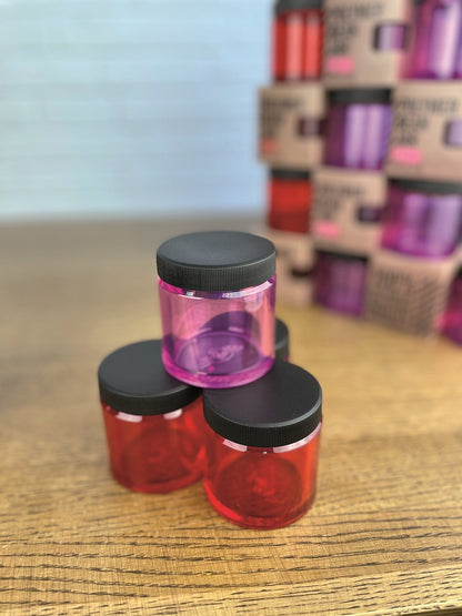 Tre farverige krukker, en pink og to røde, med sorte låg er stablet på en træoverflade. Blandt dem skiller en Comandante Polymer Bean Jar med låg i Rød sig ud. Flere krukker og kasser med lignende farver fremstår slørede i baggrunden.