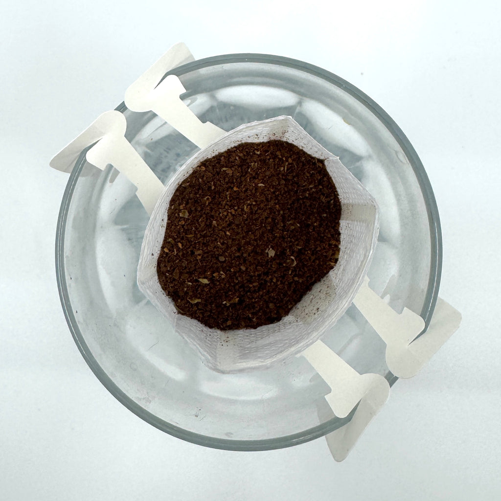 Topbillede af en glaskop med et EverBean ApS. Single Serve, No. 08 - Etiopien drypkaffefilter hængt op over kanten, indeholdende malet Arabica-kaffe, mod en hvid baggrund.