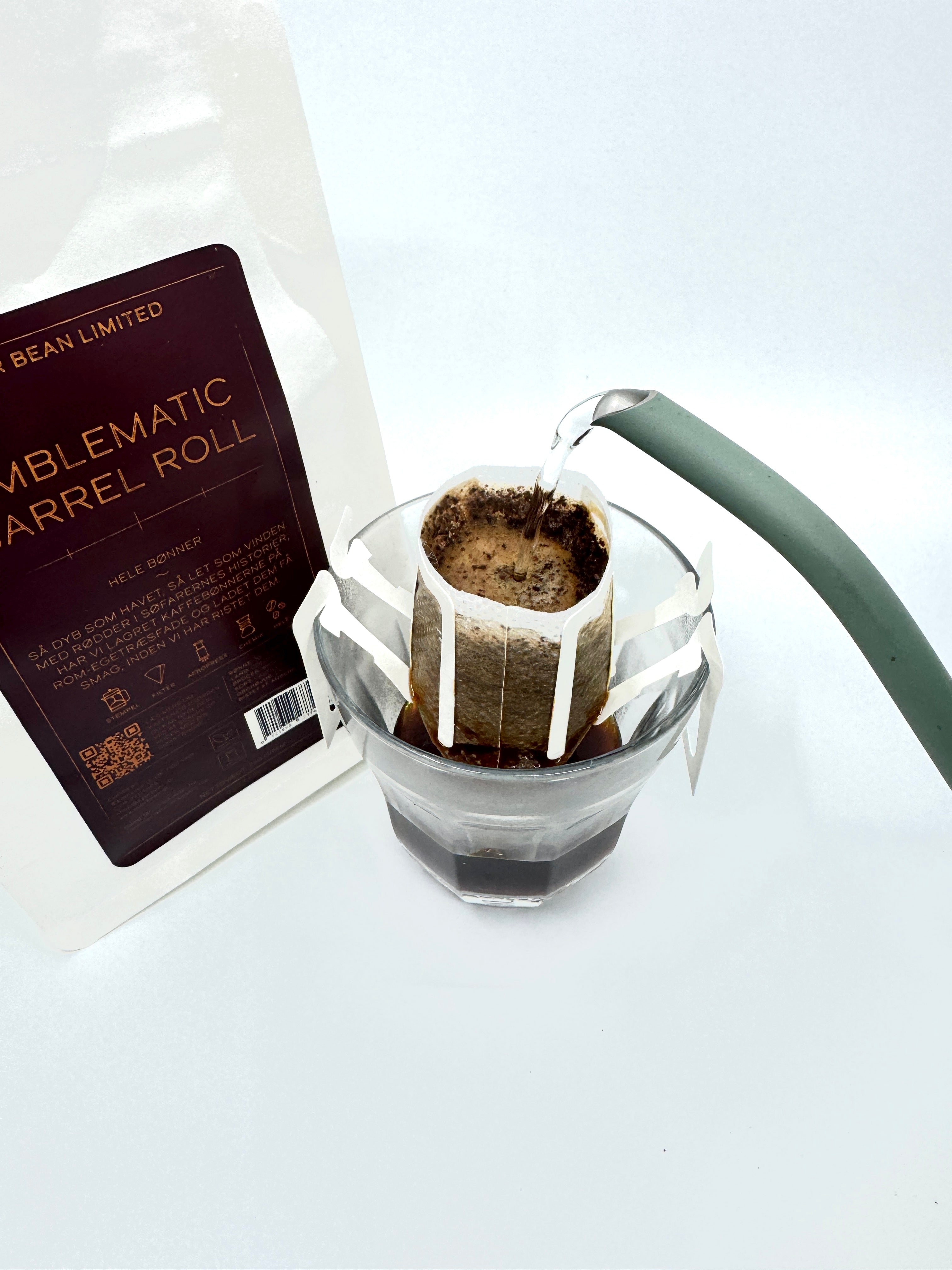 Nærbillede af en pour-over med varmt vand fra en grøn kedel på colombianske bønner i et papirfilter over glas ved siden af en hvid pose mærket "Emblematic Barrel Roll, Limited, Single Serve" fra EverBean Limited.