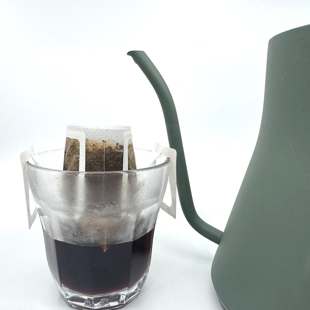 Et glas med en Single Serve, colombiansk maceration-kaffefilterpose fra EverBean Limited fastgjort til kanten, fyldt med brygget kaffe. Ved siden af står en delvis synlig grøn svanehals-kedel på en almindelig hvid baggrund.