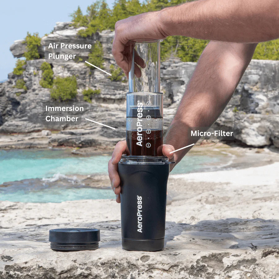 En person bruger en Aeropress Go Plus ved havet, med dens lufttryksstempel, dykkammer og mikrofilterbryggeteknologi. Grønne træer og blåt vand fremhæver scenen med en rejsetermokop ved hånden.