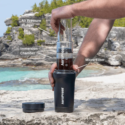 En person bruger en Aeropress Go Plus ved havet, med dens lufttryksstempel, dykkammer og mikrofilterbryggeteknologi. Grønne træer og blåt vand fremhæver scenen med en rejsetermokop ved hånden.