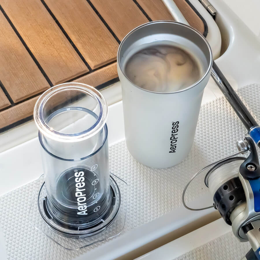 Aeropress Go Plus-kaffemaskinen fra Aeropress og et fyldt hvidt Aeropress-krus sidder på en tekstureret overflade ved siden af et fiskehjul. Kruset, med avanceret bryggeteknologi, rummer en lys drik med hvirvler. En træbaggrund står i kontrast til den hvide overflade.