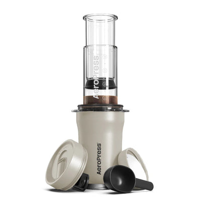 Aeropress Go Plus-kaffemaskinen fra Aeropress, med innovativ bryggeteknologi, inkluderer et stempel, målemærket kammer, to låg og en scoop - alt sammen arrangeret på en hvid baggrund. Den er perfekt kompakt til brug med den medfølgende rejsetermokop.