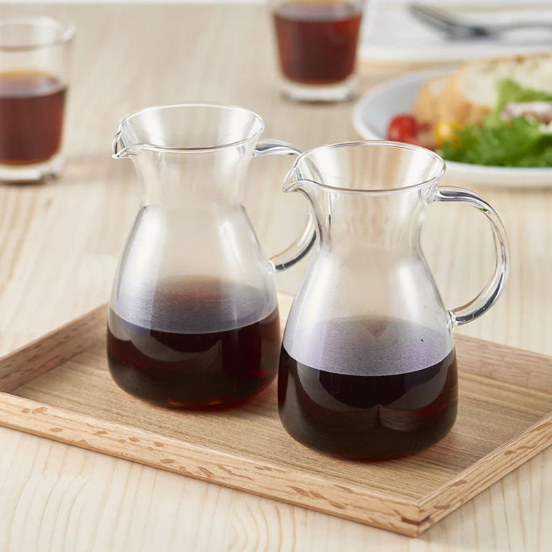 To Hario Kaffe-karafler, hver på 400 ml, fyldt med kaffe står på en træbakke på et lyst bord. I baggrunden er mad og en mørk drik sløret. Hario-karaflen holder din kaffe varm i længere tid.