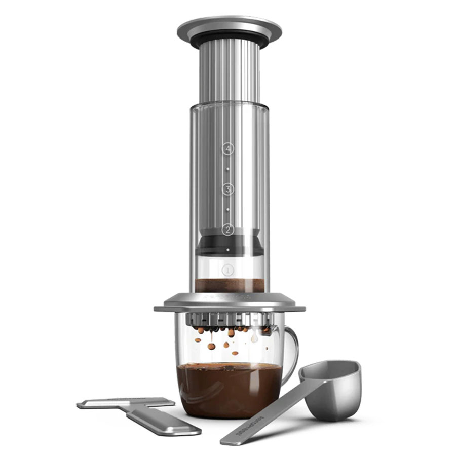 Aeropress Premium fra Aeropress brygger kaffe med 3-i-1 teknologi i en gennemsigtig cylinder af glas og rustfrit stål. Ved siden af ligger en metalske og rørepind.
