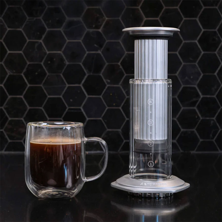 Et glaskrus med sort kaffe står ved siden af en Aeropress Premium-kaffemaskine fra Aeropress på en mørk bordplade med en sort, sekskantet flise i baggrunden.