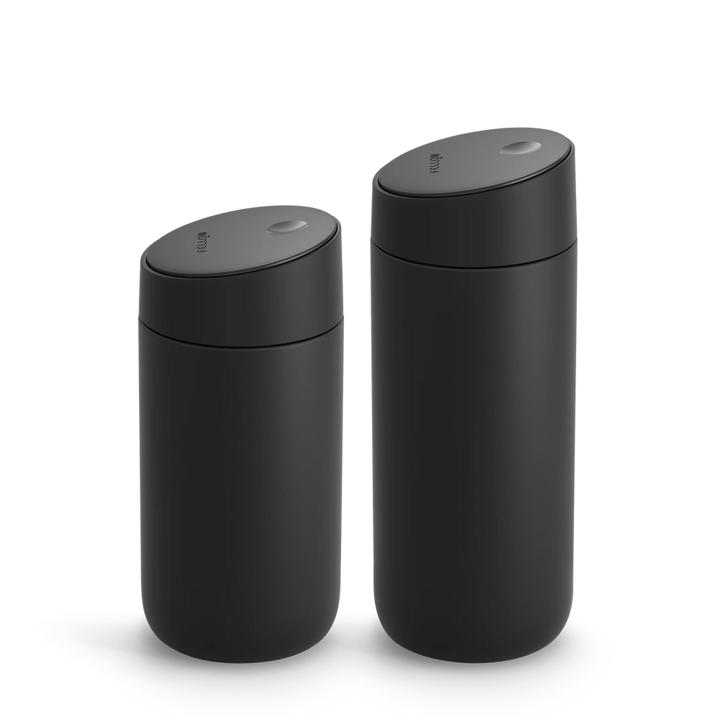 To slanke, sorte cylindriske Fellow Carter Slide-krus i forskellige højder er vist side om side. De har glatte, matte overflader og flade, ovale Slide Låg-låg med en lille knap på toppen, der afspejler det elegante design af et Carter Move-krus.