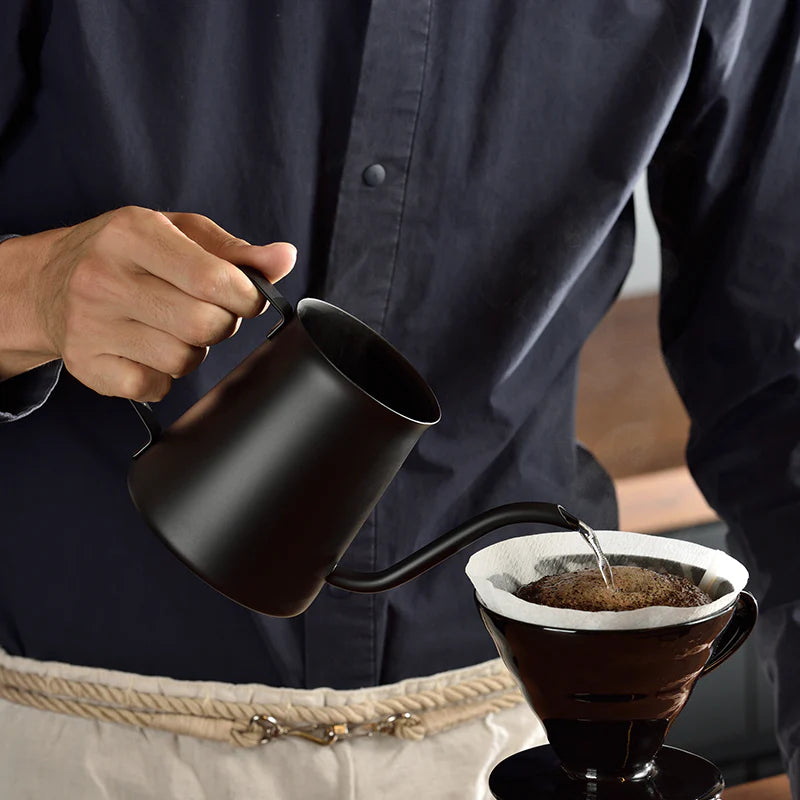 En person i mørk skjorte hælder varmt vand fra en Hario Drip-kedel, Kasuya Model, over malet kaffe i et papirfilter på en pour-over-kaffemaskine, inspireret af Tetsu Kasuyas bryggemetode.