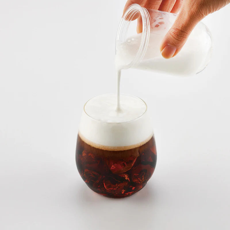 En hånd hælder cremet mælk fra Hario Latte Shaker ned i et rundt glas med iskaffe og skaber et skumlag på toppen. Scenen er sat op mod en almindelig hvid baggrund.