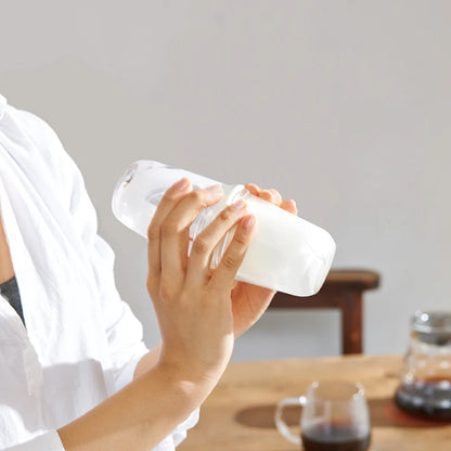 En person i hvid skjorte ryster Hario, Latte Shaker by Hario ved et træbord med en kaffekop, en karaffel og cappuccino-ingredienser i baggrunden.