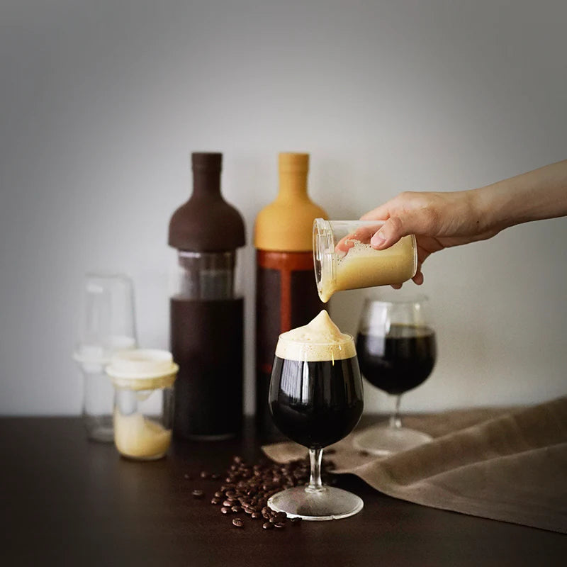 En hånd hælder cremet skum, lavet med Hario Latte Shaker, fra et glas på mørk iskaffe i et vinglas, med kaffebønner spredt på et mørkt bord og diverse flasker og krukker i baggrunden.