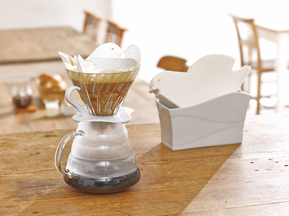 Hario keramiske pour-over kaffemaskine med et Love Birds-design sidder elegant på en glaskaraffel og brygger kaffe. I baggrunden hviler en lignende keramisk fuglefilterholder på et træbord, med sit unikke Størrelse 02 drypdesign med slørede stole bagved.