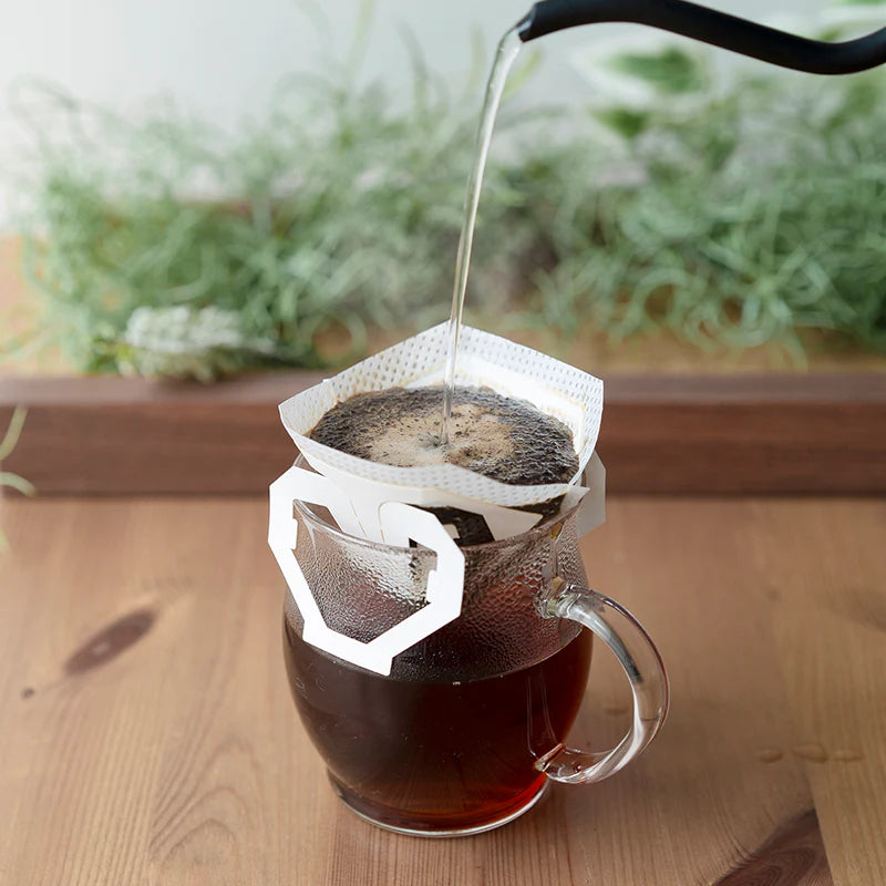 Varmt vand hældes fra en kedel ned i et Hario, en kop drypfilter, der er placeret over et krus af klart glas og brygger frisk kaffe. Opsætningen hviler på en træoverflade med grønne planter i baggrunden.