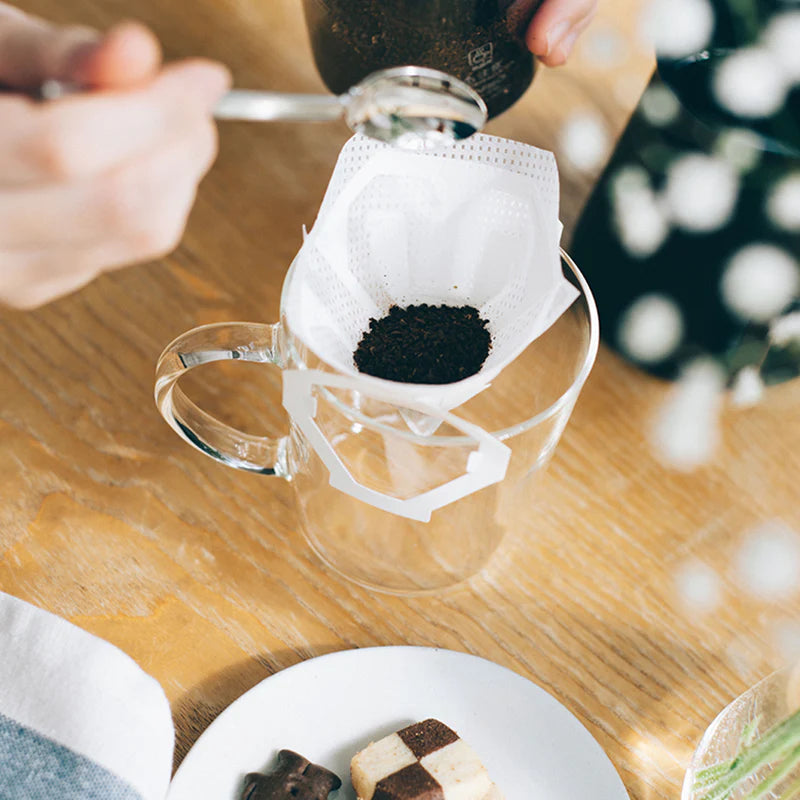 En hånd holder en ske over et glaskrus med et Hario en kop-dryppefilter, der indeholder malet kaffe. Småkager på en hvid tallerken ligger i nærheden og gør det nemt at nyde frisk kaffe overalt med Harios praktiske filter.