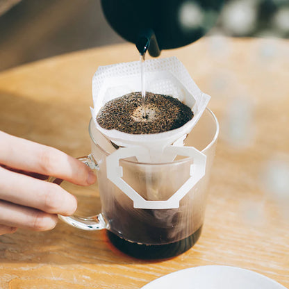 En hånd holder et glaskrus med et Hario, en kop drypfilter påsat, mens varmt vand hældes over kaffegrums i filteret. Med Hario One-Cup Drip Filter kan du brygge kaffe hvor som helst, direkte på et træbord.