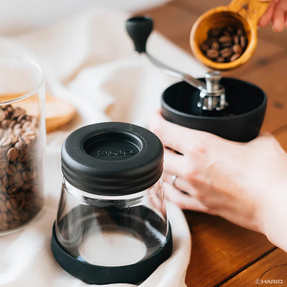 En person, der bruger en Hario, Keramisk Kaffekværn, Skerton N-kværn som kaffebønner, hældes i toppen. En glasbeholder fyldt med hele kaffebønner står i nærheden på et træbord dækket af et hvidt klæde.