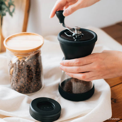 En person maler kaffebønner manuelt med en Hario Keramisk Kaffekværn, Skerton N. I nærheden sidder en krukke kaffebønner med trælåg på den stofbeklædte overflade.