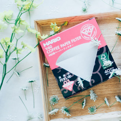 En kasse med Hario V60 papirfiltre, designet til en forbedret kaffebrygoplevelse, sidder på en træbakke omgivet af hvide og gule blomster. Den lyserøde og sorte pakke har filtrenes form og indeholder 40 "Hario, Papir Filter, Size 01" filtre.