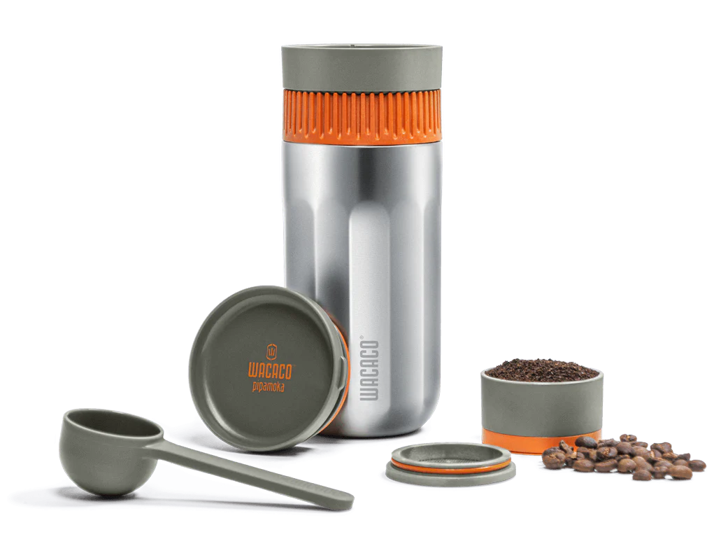 Wacaco Pipamoka fra Wacaco er en bærbar espressomaskine med en metallisk krop og orange detaljer, her vist med låg, kaffeske, beholder til malet kaffe og kaffebønner på en hvid baggrund.