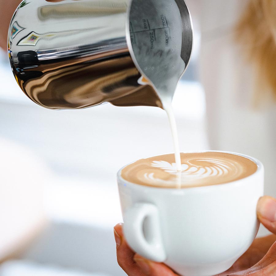 Et nærbillede af en hånd, der hælder mælk fra Rhino Coffee Gear Pro Milk Pitcher ned i en hvid kop kaffe og skaber latte art på overfladen.