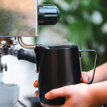 En person bruger Rhino Coffee Gear, Skummekande Stelth kande under en sølv espressomaskines dampstav. Damp stiger, mens mælken skummer mod en sløret grøn baggrund.