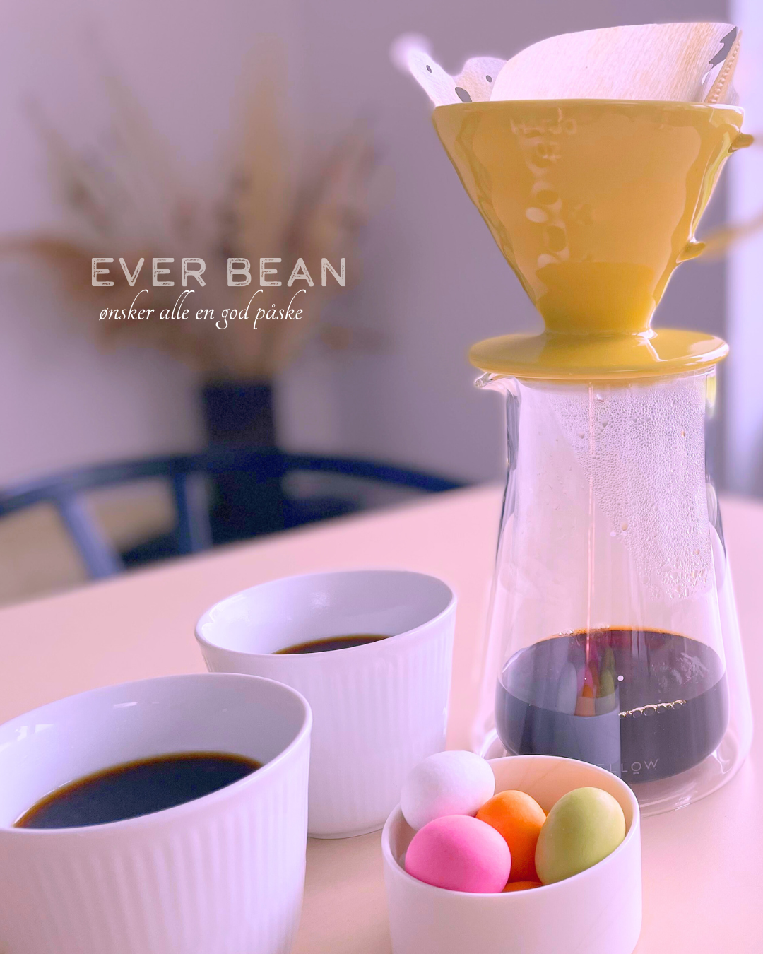 A Forårs filterpakke fra EverBean ApS. brygger kaffe i en glaskaraffel, omgivet af to hvide kopper og en skål farverige påskeæg. Teksten lyder: "EVER BEAN ønsker alle en god påske.