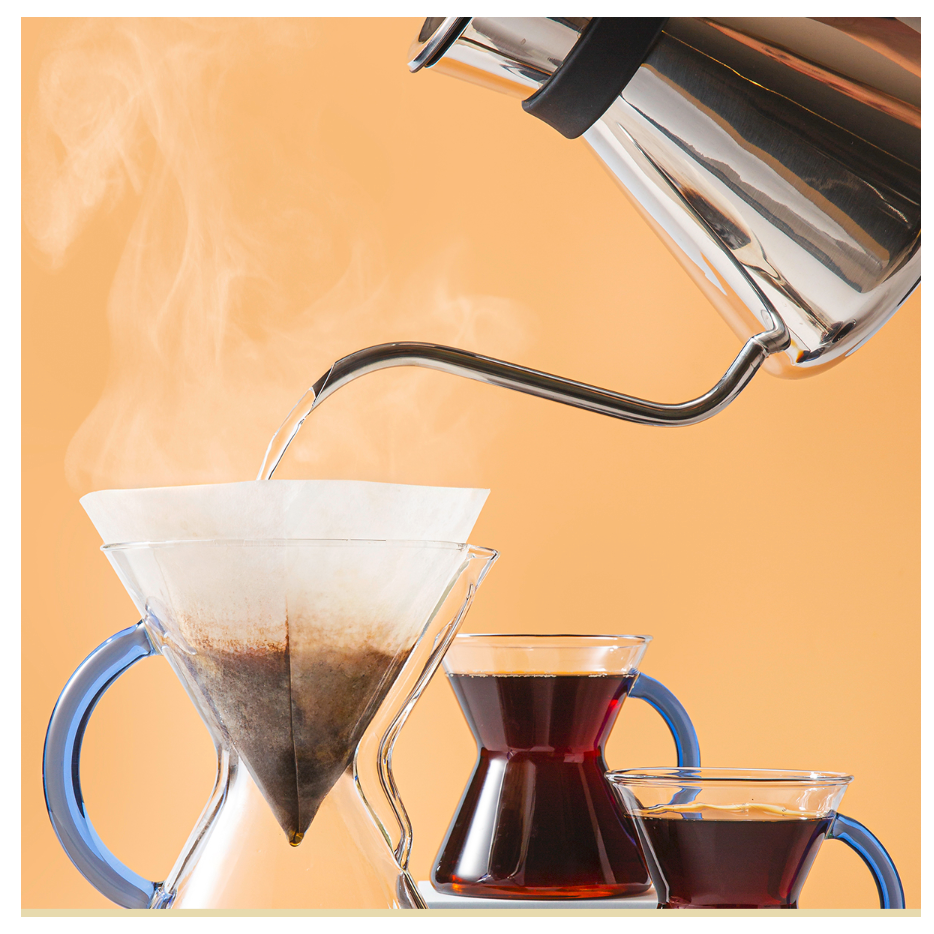 En metalkedel hælder varmt vand gennem Chemex Kaffefiltre ned i en Chemex-kaffemaskine af glas, mens dampen stiger op og to fyldte kopper står i nærheden, alt sammen på en orange baggrund.