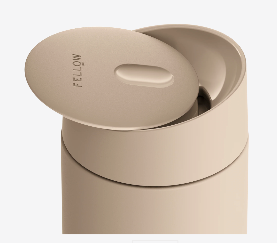 Et nærbillede af et beige Fellow Carter Slide Mug termo krus med delvist åbent låg, der viser "Fellow"-mærket på toppen og har keramisk belægning for bedre smag.