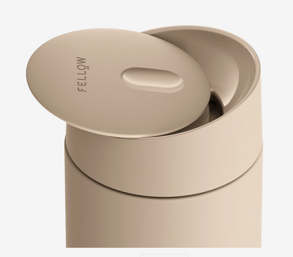 Et nærbillede af et beige Fellow Carter Slide Mug termo krus med delvist åbent låg, der viser "Fellow"-mærket på toppen og har keramisk belægning for bedre smag.