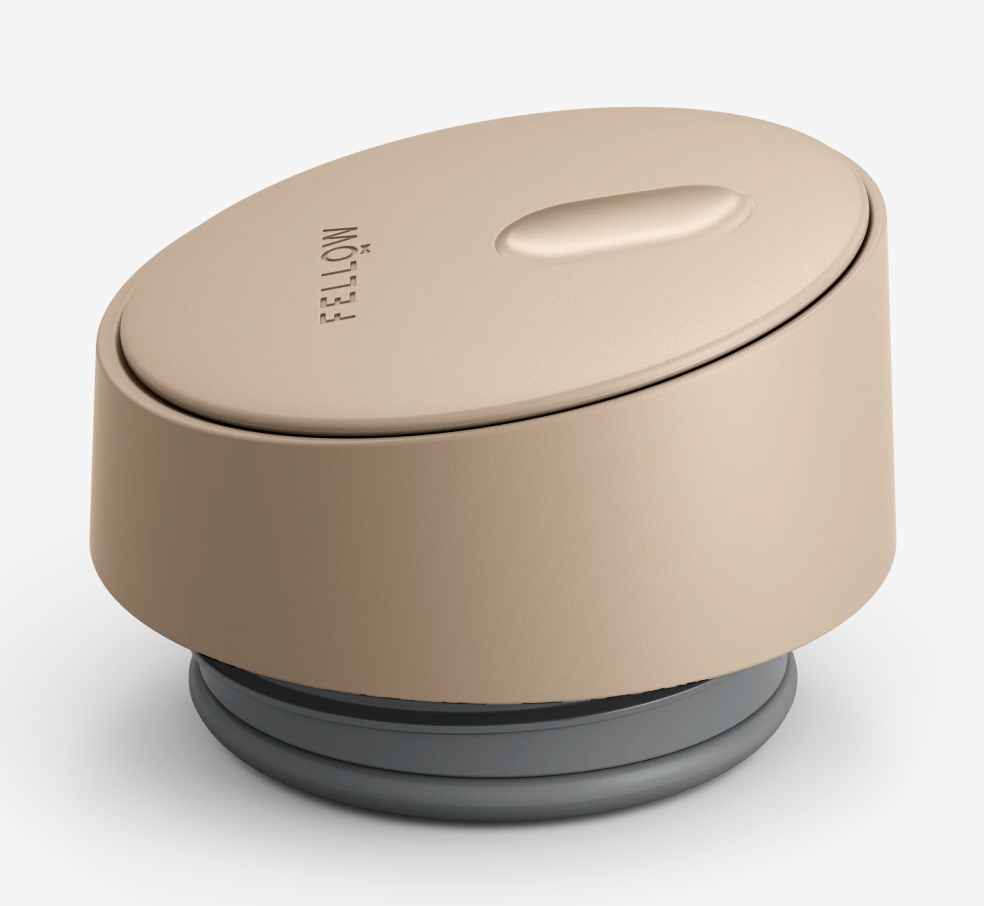 Et beige, ovalt Fellow Carter Slide Mug termo krus fra Fellow med fladt knaplåg, grå bund og keramisk belægning er vist mod en almindelig hvid baggrund.