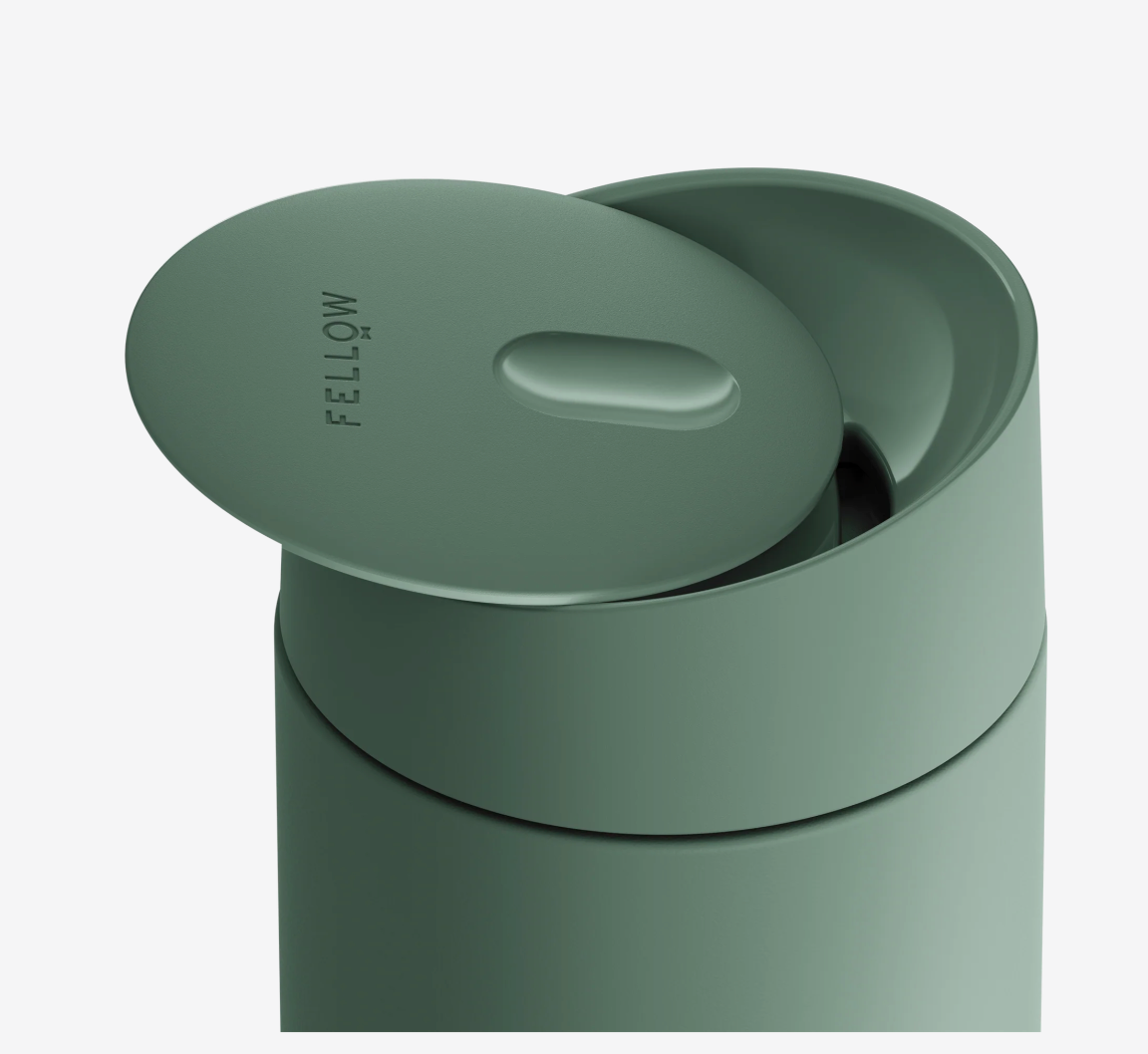 Et nærbillede af det matgrønne Fellow Carter Slide Mug, et termokrus med delvist åbent låg og et moderne, minimalistisk design. Kruset har keramisk belægning for ren smag og brandnavnet "Fellow" på toppen.