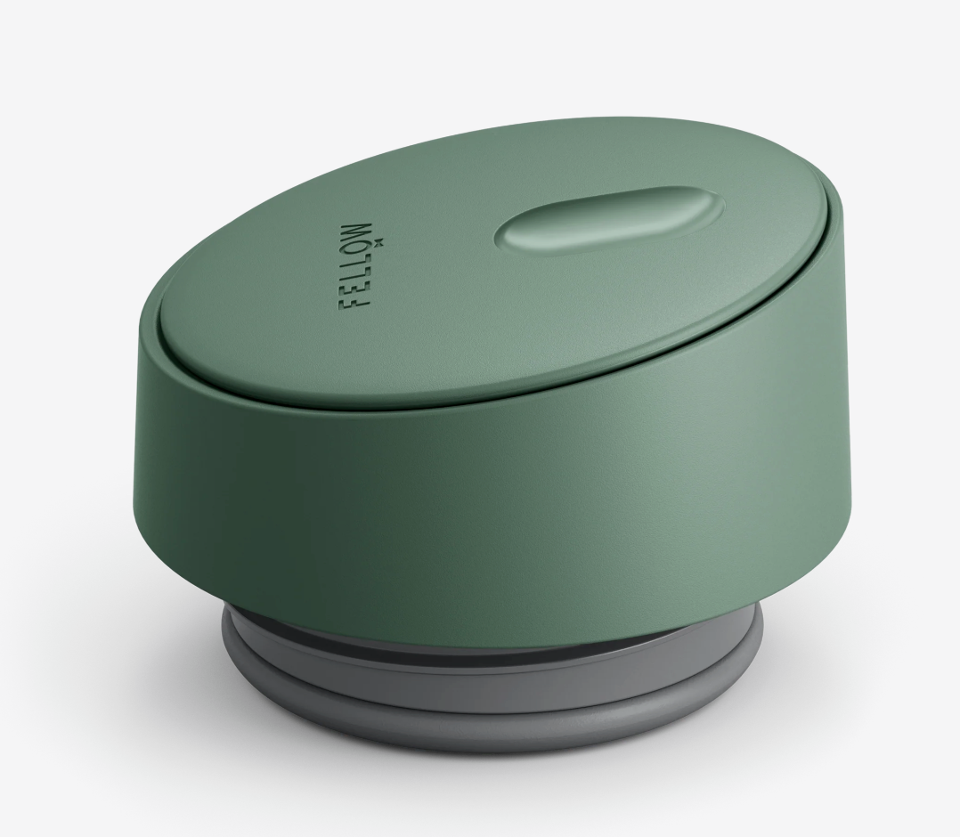 Et moderne, grønt rundt låg med en enkelt oval knap og "Fellow"-logo, designet til Fellow Carter Slide Mug Termo Krus. Det har glatte kanter, minimalistisk stil og passer perfekt sammen med en varmebevarende kopholder.