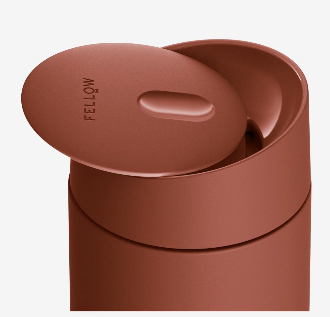 Et nærbillede af Fellow Carter Slide Mug Termo Krus i mat brun med et delvist åbent låg præget med "FELLOW". Denne varmebevarende kopholder har et moderne, minimalistisk design og holdbar keramisk belægning til ren smag.