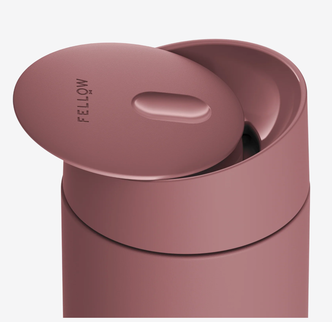 Et nærbillede af et mat rosa Fellow Carter Slide Mug, termo krus, med låget løftet for at afsløre "FELLOW" præget på toppen. Det minimalistiske design holder kaffen varm og er stilfuldt til rejser.