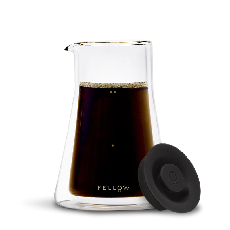 En gennemsigtig Fellow Termo Karaffel af glas fyldt med mørk kaffe har en tud på toppen og "Fellow"-etiketten i bunden. Et rundt sort låg komplementerer dets slanke design mod en almindelig sort baggrund.