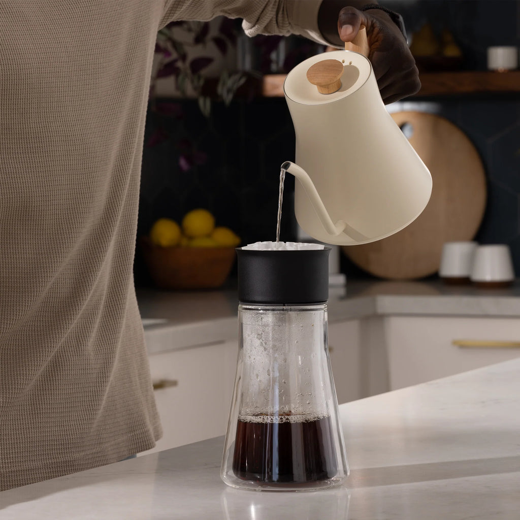 En person hælder varmt vand fra en Fellow, Stagg EKG 0,9 L elkedel med en præcisions-tud i en sort og glas kaffedryp på disken, omgivet af citroner og fade.
