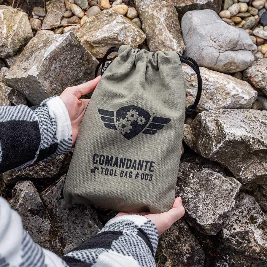 En person holder Comandante Toolbag #3 med et tandhjuls- og vingelogo mellem store grå sten - ideel til at bære din Comandante-kværn eller som en stilfuld kaffetaske.