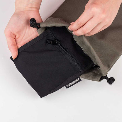 En person åbner en grøn Comandante Toolbag #3 med snoretræk og afslører en sort lynlåslomme med påskriften "BOARDMOUTH". Comandante-tasken er placeret på en hvid overflade.