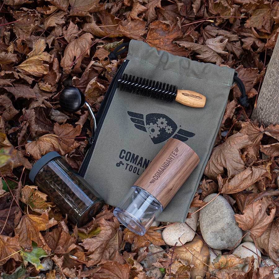 En manuel Comandante-kværn, en rensebørste og en glaskrukke ligger oven på Comandante Toolbag #3 blandt efterårsblade og sten med "COMMANDANTE TOOLS" trykt på den grønne pose.
