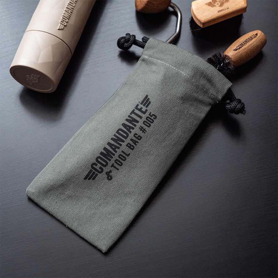 En grå Comandante Toolbag #5 fra Comandante hviler på en mørk overflade, ledsaget af værktøj med træhåndtag og en cylindrisk beholder - ideel til opbevaring af din kaffegrej.