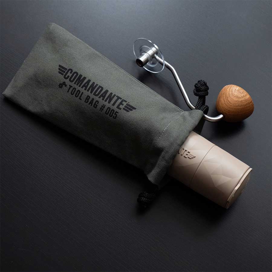 En manuel kaffekværn med træhåndtag glider delvist ud af den grønne Comandante Toolbag #5, der er perfekt til opbevaring af kaffeudstyr, og hviler på en mørk overflade.