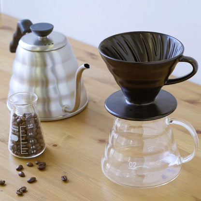 En kaffemaskine på et træbord har en sort Hario Keramisk Drip Tragt på en klar glasserver, ledsaget af en sølvfarvet svanehals-kedel. Et bæger fuld af kaffebønner og spredte bønner fuldender denne minutiøse kaffeekstraktionsscene.