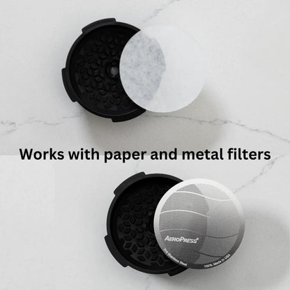 To AeroPress Flow Control-filterhætter fra Aeropress hviler elegant på en hvid marmorbaggrund - den ene holder et papirfilter, den anden et metalfilter til crema i espresso-stil. Teksten lyder: "Fungerer med papir- og metalfiltre.